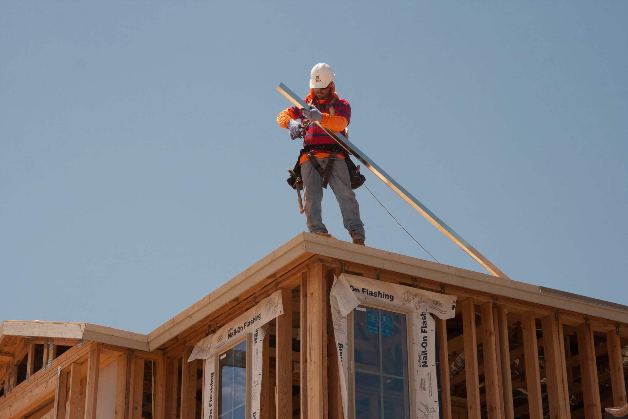 A worker in a harness standing on new wall and roof framing against a clear blue sky.